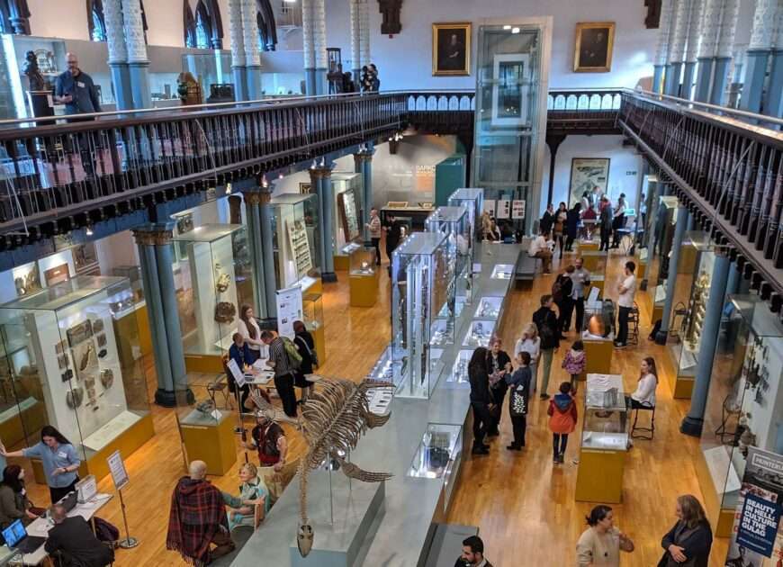 View of a museum hall from an upstairs gallery, showing groups of people looking at exhibits.